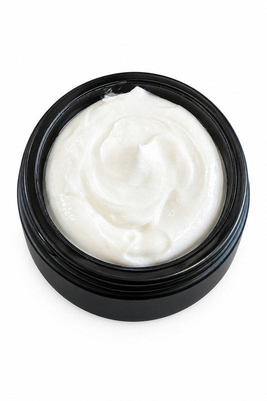 A close-up of a white cream or lotion swirled inside a round black jar, placed on a wooden tabletop with a book partially visible in the background.